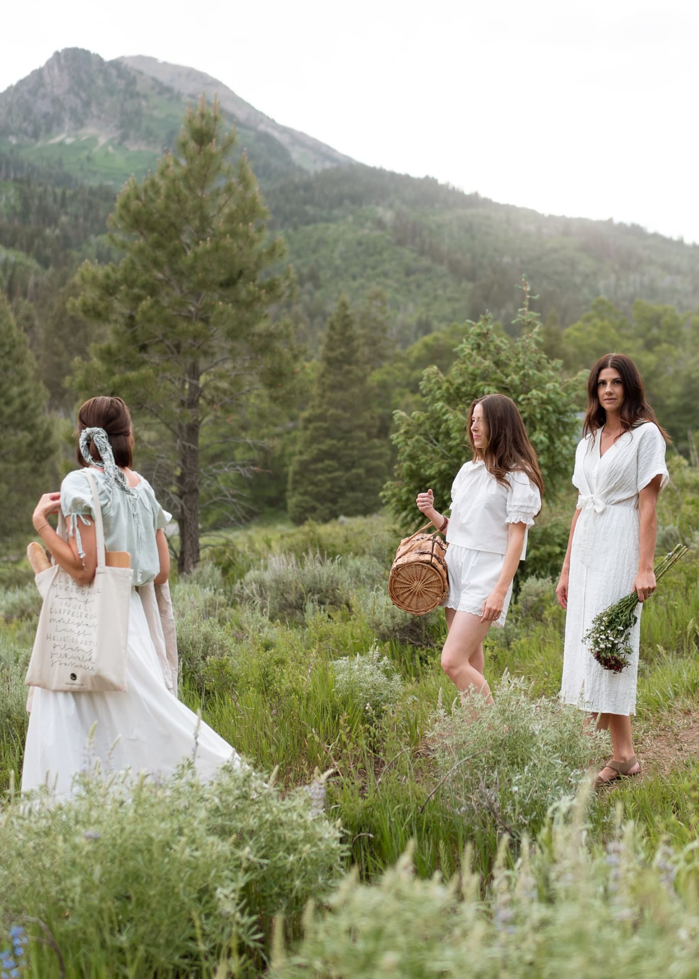 Three women in white linen walking through a mountain meadow